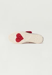 Suela de zapato de goma blanca texturizada con detalle en forma de corazón rojo. Presenta un patrón de agarre intrincado y un acento negro alrededor de los bordes.