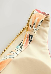 Bikini viršutinės dalies dirželio užpakalinis planas su spalvingu abstrakčiu raštu, smėlio spalvos audinys ir auksinių karoliukų apvadas palei kraštą.