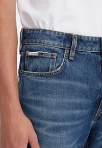 Traperice u izblijedjeloj plavoj boji, vidljivim šavovima i malom oznakom "GUESS JEANS" na prednjem džepu. Uključuje dizajn prednjeg džepa.