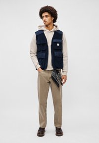 Gilet di velluto a coste blu navy con multiple tasche frontali, felpa beige con cappuccio, pantaloni khaki, bandana nera e scarpe marroni. Design semplice e funzionale.
