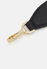 Czarna skórzana taśma z metalowym klipsem w kolorze złotym. Klips ma wygrawerowane litery. Taśma ma widoczne szwy oraz gładką fakturę.