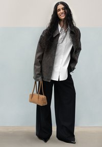 Giacca di jeans nera oversize su una camicia bianca, pantaloni neri larghi e una borsa marrone chiaro. Modella con capelli lunghi e mossi si trova contro uno sfondo chiaro.