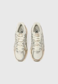 Tênis com parte superior em malha e couro em branco e cinza claro, com detalhes em bege, bico arredondado e atacadores, apresentados vistos de cima.