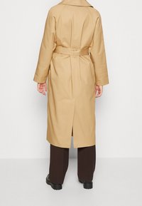 Gabardina beige con cinturón en la cintura, mangas largas y una abertura en la parte de atrás; elaborada con una tela suave. Llevada sobre pantalones oscuros con pliegues.