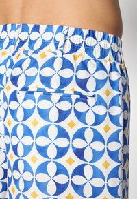 Shorts à motifs bleu et blanc avec un design géométrique comprenant des formes circulaires et des accents en losange jaunes ; comporte une poche arrière.