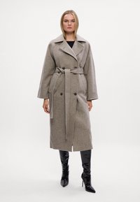 Abrigo de lana gris con cintura ceñida, presenta un cuello ancho y botones dobles. Lleva puesto sobre botas de tacón negro.