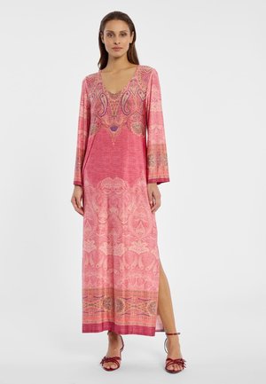 Frau trägt ein langes, rosa gemustertes Kleid mit langen Ärmeln und seitlichem Schlitz, kombiniert mit roten, hochhackigen Riemchensandalen.