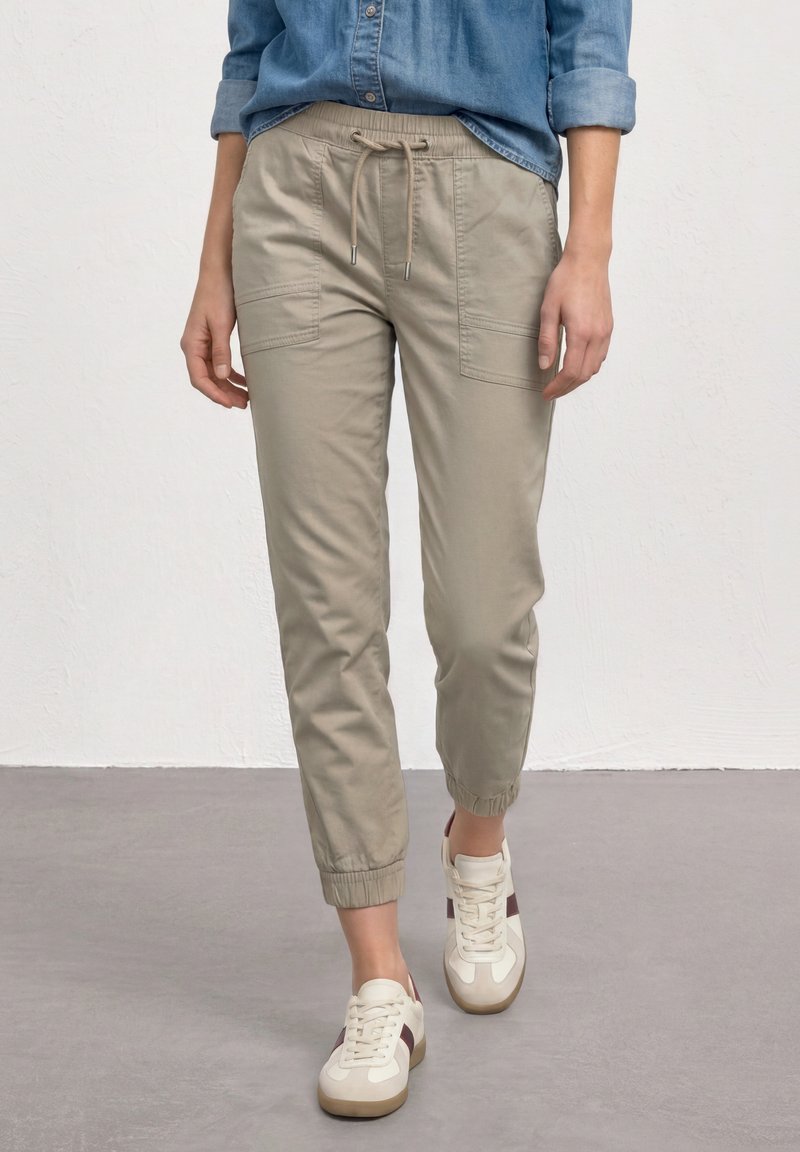 Person trägt beige Jogginghose mit Kordelzug und weiße Sneakers mit kastanienbraunen Streifen, steht auf einer grauen Fläche vor einer weißen Wand.