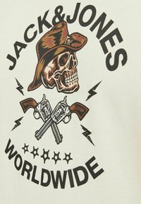 Krém színű póló, amelyen egy grafika látható, amin egy koponya barna cowboy kalapban, keresztbe tett revolverek és a "JACK & JONES WORLDWIDE" felirat szerepel.