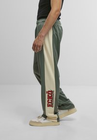 Grüne und cremefarbene Jogginghosen mit einem seitlichen Logo-Druck. Elastischer Taillenbund und glatte Textur. Kombiniert mit hellen Sneakers.