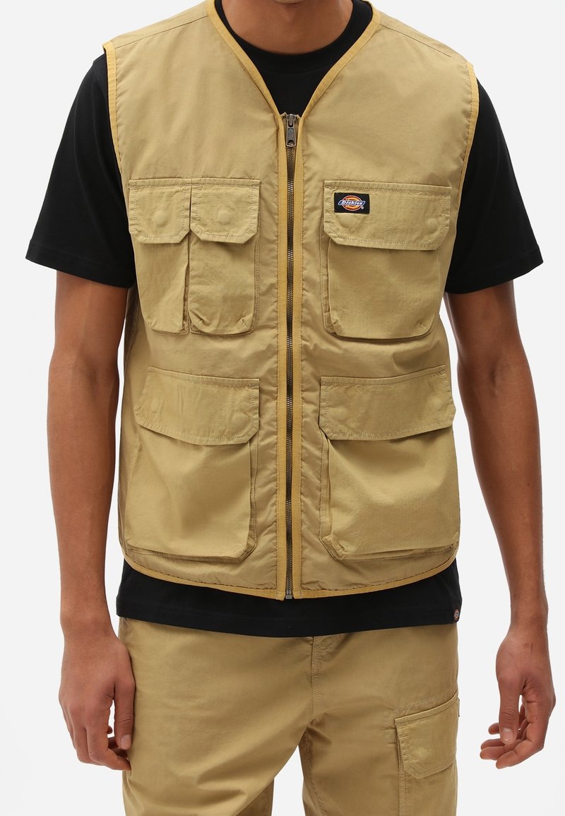 Man som bär en beige arbetsväst med flera fickor över en svart t-shirt och matchande beige cargobyxor.