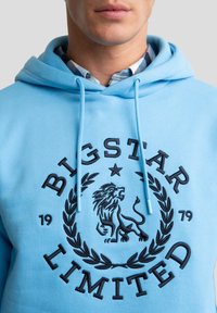 Světle modrá mikina s kapucí a šňůrkami, s černě vyšitým lvem a textem "BIGSTAR LIMITED" obklopeným designem vavřínu.