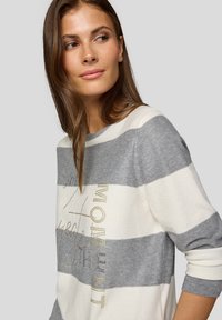 Gestreifter Pullover in Grau und Weiß, mit rundem Ausschnitt, langen Ärmeln und dekorativem Text mit metallischen Akzenten. Weiche Textur.