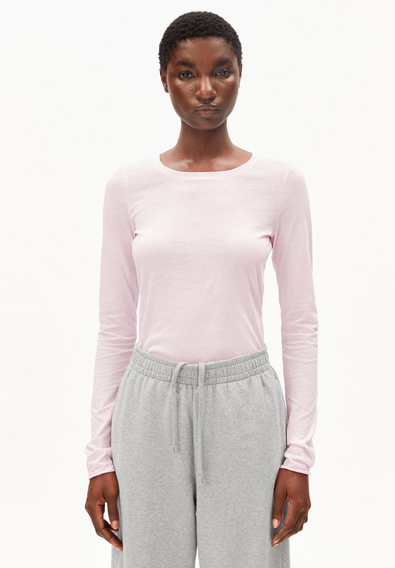 Frau mit kurzen lockigen Haaren, die ein hellrosa langärmeliges Hemd und graue Jogginghosen mit Kordelzug trägt, steht vor einem weißen Hintergrund.