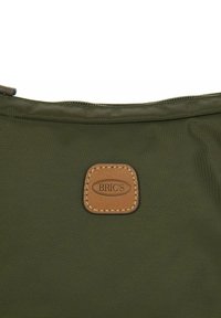 Borsa verde oliva realizzata in tessuto resistente con zip sulla parte superiore. È attaccata una toppa rettangolare in pelle marrone chiaro con la scritta "BRIC'S" in rilievo.
