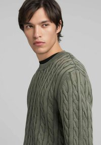 Maglione lavorato a trecce in verde oliva, con motivi testurizzati e scollo rotondo. Il tessuto è morbido con dettagli a coste nell'orlo.