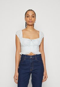 Blusa cropped azzurro chiaro con maniche a sbuffo, bordi in pizzo e dettaglio arricciato sul décolleté, abbinata a jeans scuri a vita alta.