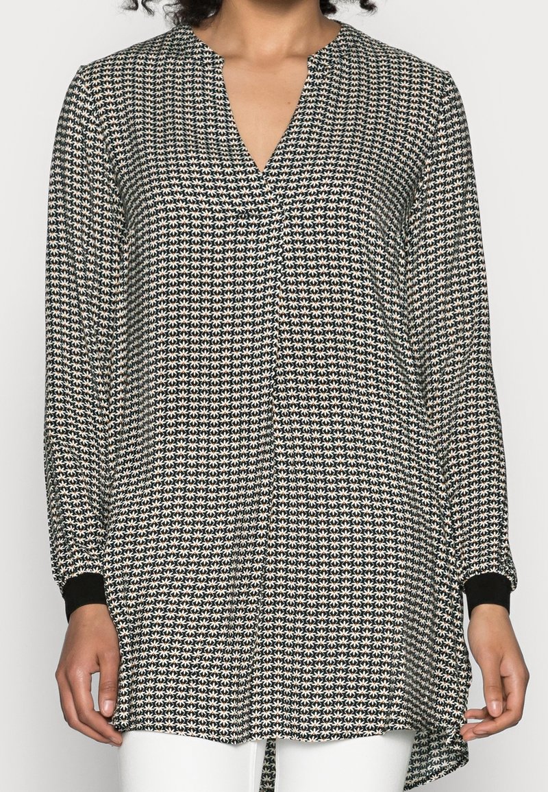 Blouse à manches longues avec un motif géométrique noir et blanc, décolleté en V et poignets noirs contrastants avec un ourlet arrière légèrement plus long.