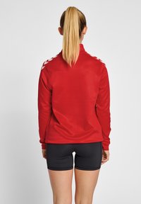 Veste de sport rouge à manches longues, col haut, avec des détails en chevrons blancs sur les épaules. Associée à un short noir. Texture lisse.