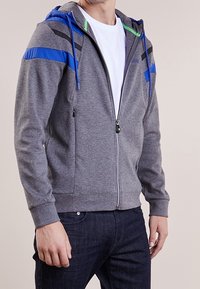 Felpa grigia con zip e accenti blu e verdi, dotata di cappuccio foderato, dettagli a strisce nere sulle spalle e tasche frontali.