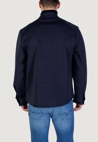 Man in een donkerblauw overhemd met lange mouwen en lichtblauwe jeans, met de rug naar de camera, tegen een effen lichte achtergrond.