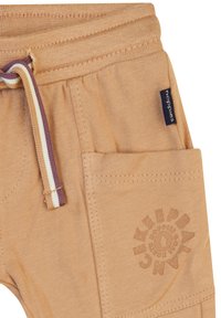 Beige Baumwollhose mit einem Kordelzug in lila und weiß, einer Seitentasche und einem geprägten Logo-Detail auf der Vorderseite.