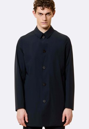 Giovane uomo con capelli ricci che indossa un cappotto nero abbottonato con colletto a punta su uno sfondo chiaro uniforme.