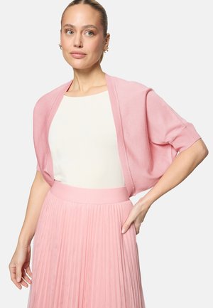 Frau trägt ein weißes Oberteil mit einem hellrosa kurzärmeligen Cardigan und einem passenden plisseeartigen rosa Rock, posiert mit einer Hand in der Hüfte.