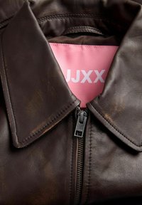 Tmavohnedá kožená bunda so zipsom vpredu a širokým golierom, zobrazujúca ružovú vnútornú etiketu s bielym nápisom "JJXX".