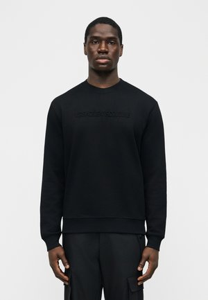 Moški stoji pred enobarvnim ozadjem, oblečen v črno jopo z reliefnim napisom in črne cargo hlače, gleda naravnost s nevtralnim izrazom.