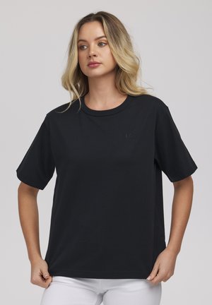 Blondynka ubrana w luźną czarną koszulkę z krótkim rękawem i białe spodnie, trzyma za brzeg koszulki, patrzy lekko w prawo.