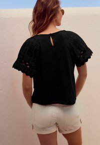 Czarna bluzka z fakturowanymi, falbaniastymi rękawami, z detalem w kształcie dziurki na plecach. Noszona z białymi szortami. Prosty, casualowy design.