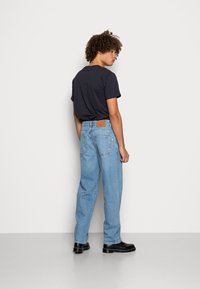 Jeans de ganga azuis com um corte descontraído, apresentando um remendo castanho na cintura, acompanhados por uma t-shirt preta de mangas curtas e botas pretas.