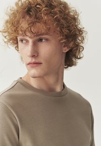 Tricou bej cu mânecă scurtă și decolteu rotund, realizat dintr-un material neted. Modelul are părul creț până la umeri și o cercei subtili.