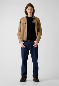 Chemise marron à boutons portée sur un t-shirt noir, associée à un jean bleu et des bottes foncées. La chemise possède une poche sur la poitrine et une coupe classique.