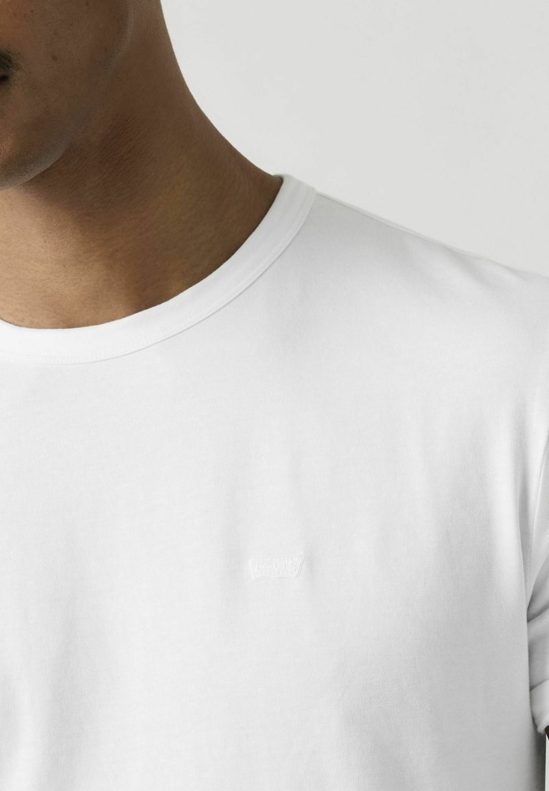 Primo piano di una persona che indossa una maglietta bianca semplice con scollo a girocollo e un logo in rilievo sottile sul petto.