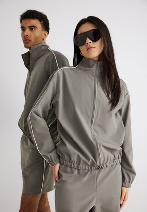 Dva modela nose sivi športni džemperi s bijelim detaljima; ženski model u hlačama i sunčanim naočalama stoji naprijed, muški model u kratkim hlačama stoji iza.