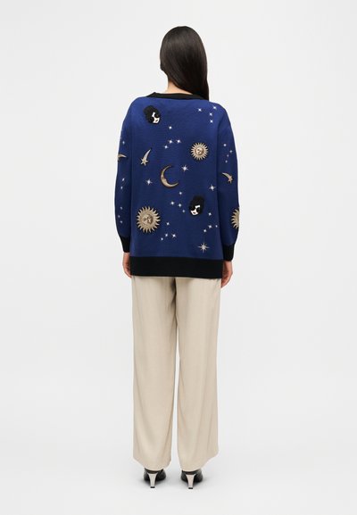 Pull en tricot bleu avec broderies célestes, comprenant des motifs de lunes, étoiles et soleils en or et noir, assorti à un pantalon beige à jambes larges.