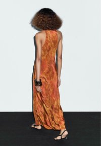 Robe maxi en orange brûlé avec un motif floral texturé. Elle possède un col halter, une forme fluide et est assortie de sandales noires et de bracelets.