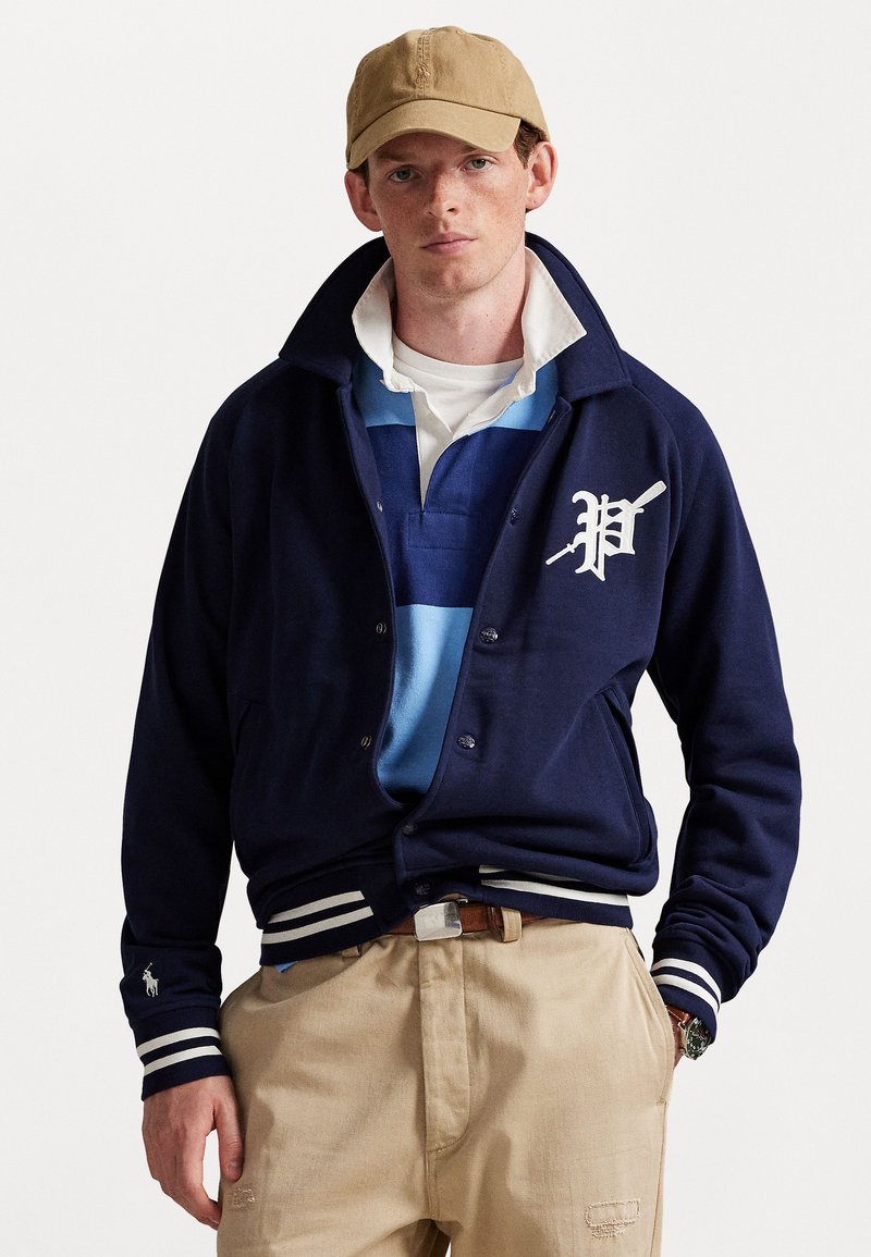 Giovane uomo che indossa una giacca varsity blu scuro con strisce bianche, una maglietta da rugby blu, pantaloni beige, un cappellino color sabbia e un orologio da polso, in posa davanti a uno sfondo neutro.