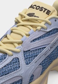 Κοντινό πλάνο σε μπλε πλεκτό αθλητικό παπούτσι Lacoste με κρεμ κορδόνια και γλώσσα, με κεντημένο λογότυπο και το κείμενο «Από το γήπεδο στους δρόμους από το 1933».