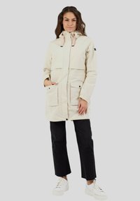 Veste d'extérieur beige avec une capuche, fermetures à boutons et à pression, deux grandes poches avant et accents rose pâle sur la capuche.