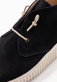 Černé semišové boty s texturovaným povrchem, smetanovými tkaničkami a silnou bílou gumovou podrážkou se svislými rýhami.