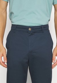 Marineblå chinos med flad front, en knaplukning, sidelommer og en glat tekstur. Båret med en lyseblå tætsiddende T-shirt.