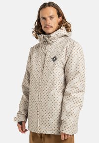 Mężczyzna z kręconymi włosami do ramion, ubrany w beżową kurtkę z kapturem w wzory i brązowe spodnie, stoi na jasnym tle.