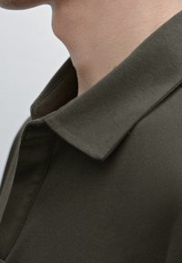 Chemise vert olive foncé avec un col rabattu, présentant une texture lisse et mate ainsi qu'un design minimaliste, mettant en avant des lignes épurées et la simplicité.