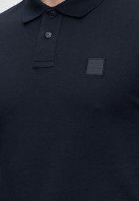 Close-up van een persoon die een donker marineblauw poloshirt draagt met een sluiting met twee knopen en een vierkante logopatch met de letters "BOSS" op de borst.