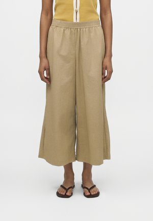 Du milieu du corps jusqu'aux pieds d'une personne portant un pantalon large beige et court, un haut jaune sans manches, et des sandales marron type tongs, debout sur fond blanc.