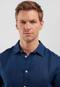 Camicia in lino blu navy con colletto, bottoni frontali e una trama sottile. Presenta dettagli bianchi sui bottoni per offrire contrasto.