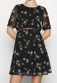 Mekko mustasta kukallisesta läpikuultavasta kankaasta, jossa on lyhyet hihat, pyöreä pääntie ja punottu ruskea vyö vyötäröllä sekä laajahelma.